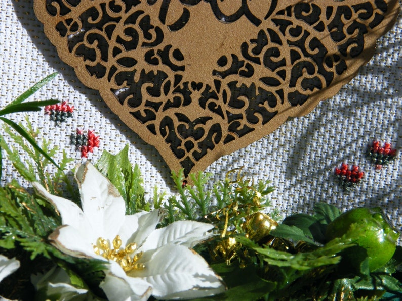 Pode incluir: Uma flor de poinsettia branca com detalhes dourados, cercada por folhagem verde e um ornamento em forma de cora&ccedil;&atilde;o de madeira marrom esculpido.