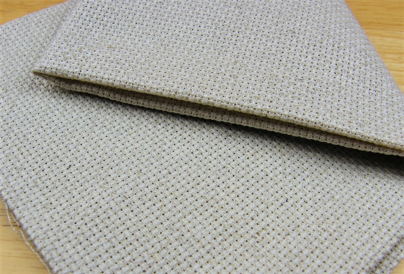 May include: Close-up of two pieces of beige cross-stitch fabric. The fabric has a woven texture with small, evenly spaced holes, ideal for needlework. The edges are neatly finished, suggesting a craft project.