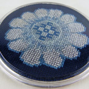 Può includere: Un sottobicchiere rotondo con un bordo in plastica trasparente, decorato con un motivo floreale ricamato in blu e bianco.