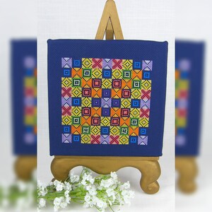 Peut inclure: Un motif de point de croix coloré sur une toile bleue, monté sur un chevalet en bois. Le motif présente un motif répétitif de carrés de différentes couleurs, notamment jaune, orange, rouge, violet et vert.