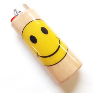 May include: A wooden lighter with a yellow smiley face design. The lighter is a standard size and has a red plastic top.