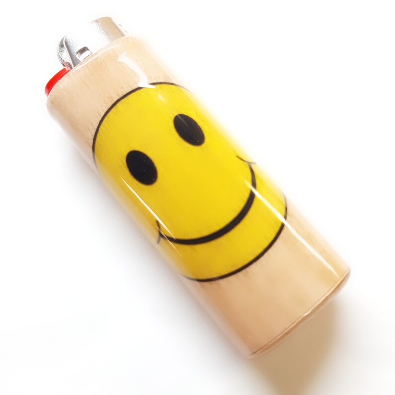 May include: A wooden lighter with a yellow smiley face design. The lighter is a standard size and has a red plastic top.