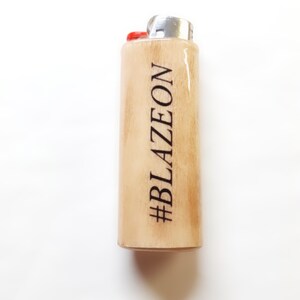 以下が含まれることがあります： 銀色のトップと赤いボタンが付いた木製のライター。ライターには「#BLAZEON」という文字が刻まれています。