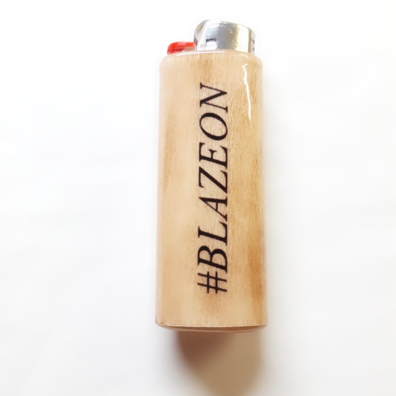 以下が含まれることがあります： 銀色のトップと赤いボタンが付いた木製のライター。ライターには「#BLAZEON」という文字が刻まれています。