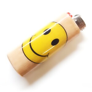 May include: A wooden lighter with a yellow smiley face design. The lighter has a silver top with the text "Zippo" and "Made in USA" printed on it.