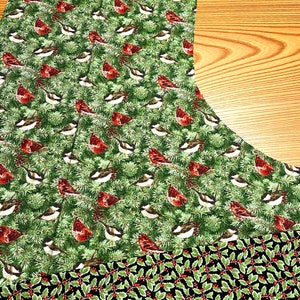 May include: A green fabric with a repeating pattern of red cardinals and pine branches. The bottom portion of the fabric is black with a repeating pattern of green holly leaves and red berries.