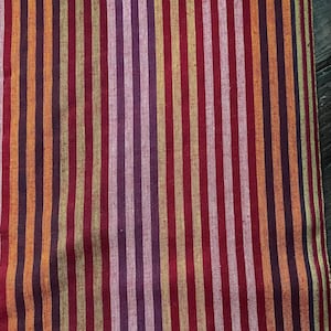 May include: A woven fabric featuring a repeating vertical striped pattern in shades of deep burgundy, vibrant orange, muted gold, and soft pink. The textile displays a consistent linear design with varying stripe widths that run parallel across the surface.