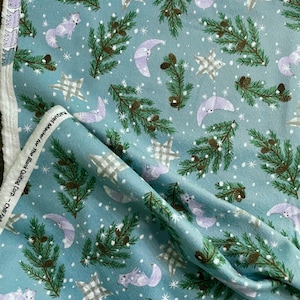 May include: A light blue fabric with a winter-themed pattern. The design includes white snowflakes, gray plaid stars, purple crescent moons, and purple foxes. Green pine branches with brown pinecones complete the design. The fabric is folded.