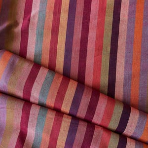 May include: A textile fabric featuring a repeating vertical stripe pattern in a vibrant color palette of magenta, deep purple, burnt orange, soft pink, and muted teal. The material has a visible woven texture with a slight sheen across the surface.