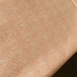 May include: A close-up of a peach-colored fabric with a subtle white pattern. The fabric has a textured, woven appearance.