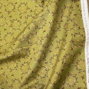 May include: Green fabric with a repeating pattern of circular wreaths, stars, and floral accents. The wreaths are brown with yellow and red details, set against a light green background. The fabric is slightly wrinkled.