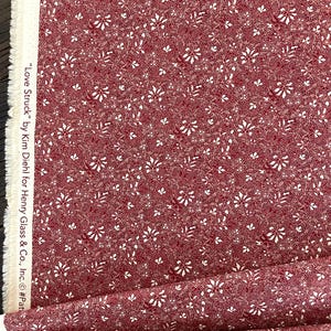 May include: Red fabric with a white floral pattern. The fabric is printed with small white flowers and leaves on a red background. The text "Love Struck" by Kim Diehl for Henry Glass & Co., Inc. is printed on the edge of the fabric.