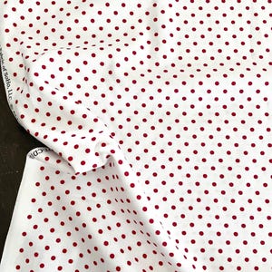 May include: White fabric with a repeating pattern of evenly spaced, small, red polka dots. The fabric is folded and draped, showing the texture and flow of the material. The fabric is likely cotton or a similar material.