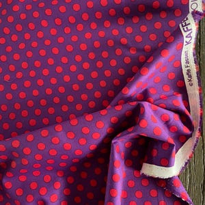 May include: A vibrant purple fabric with a repeating pattern of large, red polka dots. The fabric is folded, showing the texture and sheen. The selvedge edge displays the text "Kaffe Fassett" and "COLLECTION".