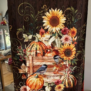 May include: A brown fabric panel with a fall-themed design. The panel features a wooden crate filled with pumpkins, sunflowers, and birds. The background is a dark brown woodgrain pattern with swirls and vines.