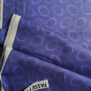 May include: A folded piece of fabric with a repeating pattern of large, blue-purple ovals filled with tiny black dots. The fabric has a white selvage edge with text that reads "FASSETT CO." and "Free Spirit".