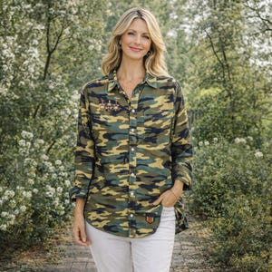May include: A camouflage-patterned button-down shirt in shades of green, tan, and navy blue. The shirt has a chest pocket, a small patch near the hem, and a decorative pin. The model is wearing white pants.