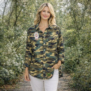 May include: A camouflage-patterned button-down shirt with a colorful skull patch on the chest and a green embroidered design near the hem. The shirt is in shades of green, brown, and tan, paired with white pants. The shirt has long sleeves and a relaxed fit.