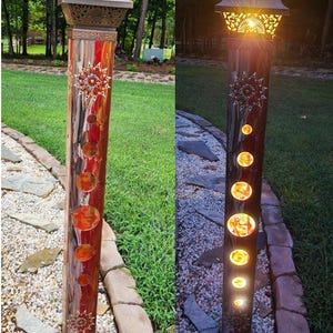 Pode incluir: Uma luz de jardim solar com uma parte superior de metal decorativa e uma coluna de madeira com um design de mosaico. A luz está acesa na segunda imagem, mostrando o brilho quente das lâmpadas.