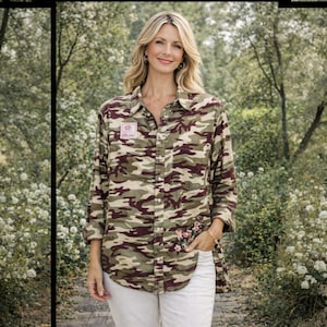 May include: A camouflage-patterned button-down shirt in shades of green, brown, and tan. The shirt features a small floral embroidery and a postage stamp patch. The model is wearing white pants.
