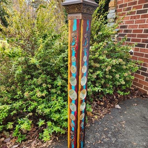May include: A tall, decorative wooden post with a bronze-colored top. The post is painted with vertical stripes of turquoise, yellow, red, and brown. It features decorative cutouts and embedded, clear, circular accents. The post is outdoors, near greenery.