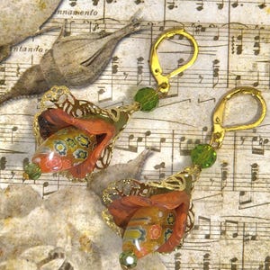 May include: A pair of handcrafted earrings featuring a floral design. The earrings have gold-colored ear wires, green beads, and orange, yellow, and blue floral accents. The earrings are set against a background of sheet music.