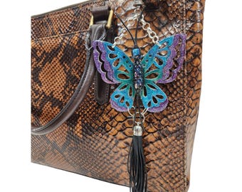 May include: A brown snakeskin-patterned handbag with a decorative butterfly charm. The butterfly charm is blue and purple with a black tassel and silver chain. The handbag has a brown leather handle and gold hardware.