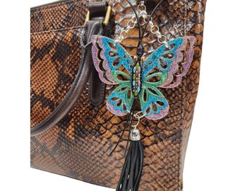 May include: A brown snakeskin-patterned handbag with a decorative butterfly charm. The butterfly is blue, green, and purple with a black tassel. The bag has a brown handle and gold-colored hardware.