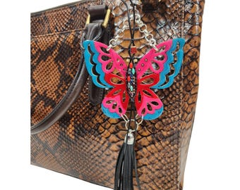 May include: A decorative butterfly charm with pink and blue wings, embellished with colorful gemstones, hangs from a brown snakeskin-patterned handbag. The charm has a black tassel and silver chain.