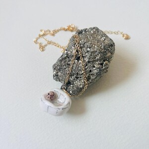 May include: A gold chain necklace with a white pendant. The pendant is a small, white ceramic cradle with a baby inside. The necklace is displayed on a rough, gray rock. The chain is a delicate design.