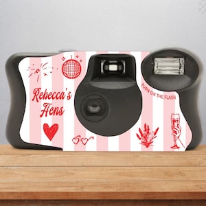 May include: A black disposable camera with a pink and white striped design. The camera has a flash and a lens cover. The design includes red illustrations of a disco ball, fireworks, a heart, sunglasses, a glass of champagne, and a sprig of lavender. The text "Rebecca's Hens" is printed on the camera.