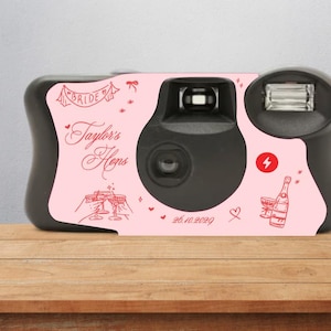 May include: Black disposable camera with a pink overlay. The overlay features the text "Bride" and "Taylor's Hens" in red script, along with illustrations of champagne glasses and a bottle. The date "26.10.2020" is also printed on the camera.