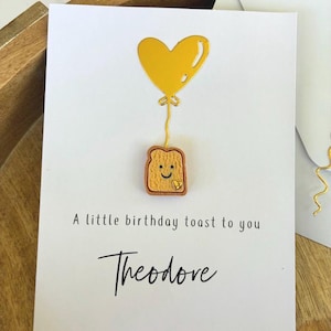 Puede incluir: Tarjeta de felicitación blanca con un globo en forma de corazón amarillo y una ilustración de una tostada sonriente. El texto "A little birthday toast to you" y el nombre "Theodore" están escritos en cursiva.