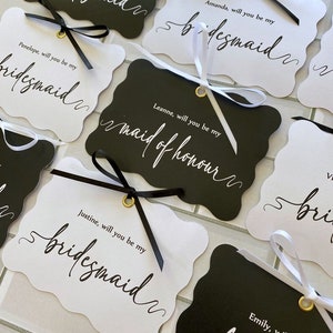 May include: Scalloped-edge tags in black and white with gold eyelets and tied ribbons. The tags feature elegant calligraphy that reads "Anastashia, will you be my bridesmaid", "Amanda, will you be my bridesmaid", and other names.