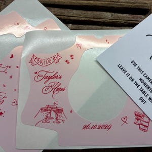 Puede incluir: Adhesivo rosa claro para c&aacute;mara con ilustraciones de l&iacute;nea roja de copas de champ&aacute;n, corazones, estrellas y el texto "Taylor's Hens" y "26.10.2029". Una tarjeta de instrucciones blanca es parcialmente visible.