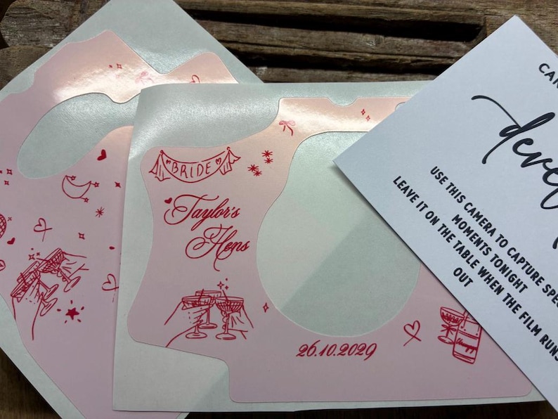 Puede incluir: Adhesivo rosa claro para c&aacute;mara con ilustraciones de l&iacute;nea roja de copas de champ&aacute;n, corazones, estrellas y el texto "Taylor's Hens" y "26.10.2029". Una tarjeta de instrucciones blanca es parcialmente visible.