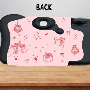 Puede incluir: Una c&aacute;mara desechable negra con un panel frontal rosa claro que presenta ilustraciones de l&iacute;nea roja de una bola de discoteca, corazones, una luna creciente, copas de champ&aacute;n, un regalo, un pastel y estrellas. La palabra BACK est&aacute; impresa arriba.