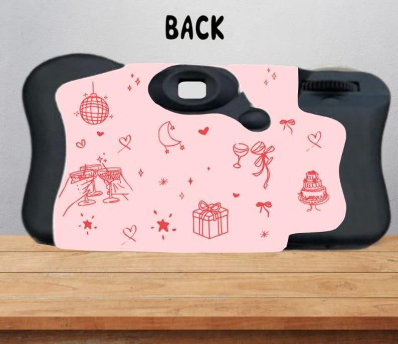 Puede incluir: Una c&aacute;mara desechable negra con un panel frontal rosa claro que presenta ilustraciones de l&iacute;nea roja de una bola de discoteca, corazones, una luna creciente, copas de champ&aacute;n, un regalo, un pastel y estrellas. La palabra BACK est&aacute; impresa arriba.