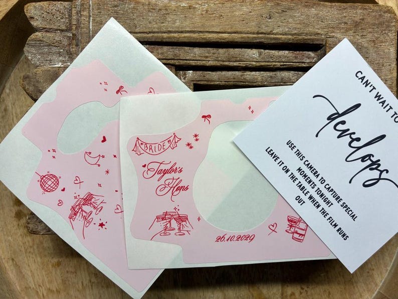 Puede incluir: Una funda de c&aacute;mara rosa claro con dibujos lineales rojos de una bola de discoteca, copas de champ&aacute;n, corazones y estrellas. El texto dice Taylor's Hens, Bride y 26.10.2029. Una tarjeta blanca adjunta dice Can't wait to see how it develops.