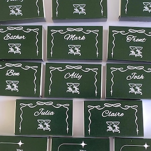 Puede incluir: Cajas de cerillas verde oscuro con nombres blancos e ilustraciones de copas de champán. Algunas cajas muestran el texto "The Perfect Match".