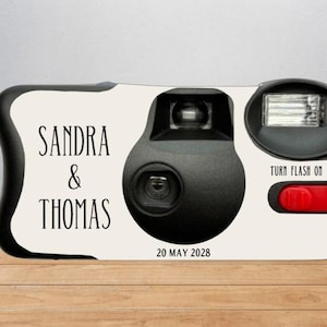 Puede incluir: Una cámara desechable con exterior blanco que presenta el texto negro SANDRA & THOMAS y 20 MAY 2028. La cámara tiene una carcasa de lente central negra, un flash negro y un botón rojo con la etiqueta TURN FLASH ON en texto negro.