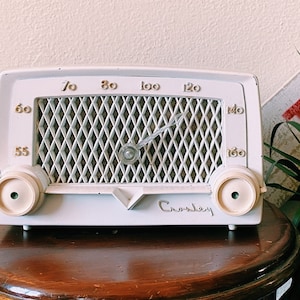 May include: A vintage white Crosley radio with gold numbers and a dial. The radio features a diamond-patterned speaker cover and two cream knobs. The radio sits on a dark wooden table. A sign with red text is visible in the background.