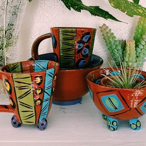 Puede incluir: Una colección de tazas de cerámica pintadas a mano y una pequeña jardinera. Las tazas presentan diseños geométricos en tonos rojos, verdes, azules y amarillos. Una taza tiene pequeños pies azules. Un jarrón de cristal transparente y vegetación en el fondo.