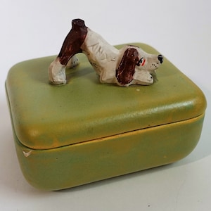 May include: A green ceramic trinket box with a white and brown ceramic dog figurine on top. The dog is in a playful pose with its head tilted to the side.