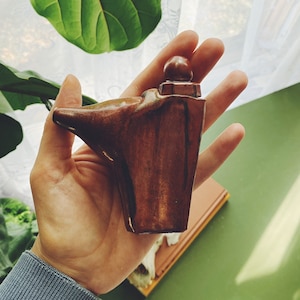 May include: A hand is holding a small, wooden, brown and black figurine. The figurine is shaped like a person with a rounded head and a long, flowing robe. The figurine is standing on a small, wooden base.