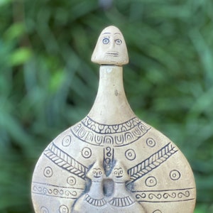May include: A beige ceramic bottle with a decorative design. The bottle features a stopper with a face, and the body has an embossed pattern with two figures. The design includes geometric shapes and lines, creating a unique, handcrafted look.