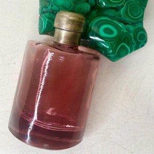 Puede incluir: Una botella de vidrio vintage con una tapa plateada y un líquido rosa en su interior. La botella está sobre una superficie blanca con una piedra de malaquita verde detrás.