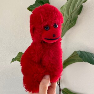 May include: A bright red plush monkey toy with black eyes and a black stitched smile. The monkey is held in a hand against a white background with green leaves.