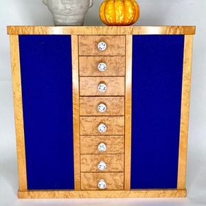 May include: A wooden jewelry box with seven drawers and blue velvet lining. The drawers have white porcelain knobs with a blue floral design. The box is sitting on a white surface with a potted plant and a small pumpkin.