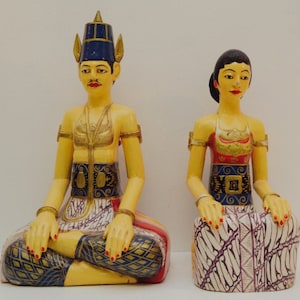 Pode incluir: Duas estátuas de madeira pintadas à mão de um homem e uma mulher em trajes tradicionais indonésios. O homem usa um cocar azul e detalhes dourados. A mulher tem um top vermelho e dourado e uma saia estampada. Ambos estão sentados.